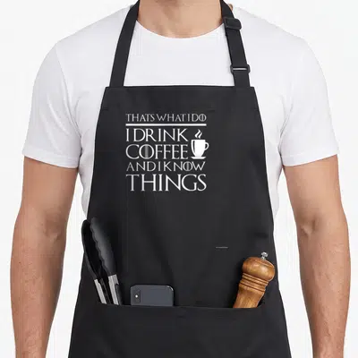 Funny drink coffee apron - White stacked text THATS WHAT I DO I DRINK COFFEE AND I KNOW THINGS with small steaming cup — shown on a black cotton canvas apron against a white background