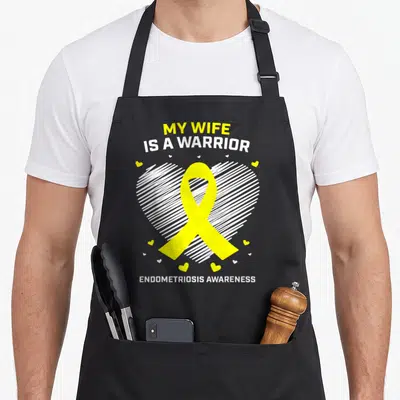 Endo husband wife aprons - My Wife Is A Warrior text with yellow ribbon over white hatched heart — shown on a black cotton canvas apron against a white background