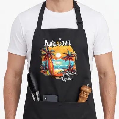 Dominican family aprons - Punta Cana beach scene with palm trees, summer surfboard, and Dominican Republic text — shown on a black cotton canvas apron against a white background