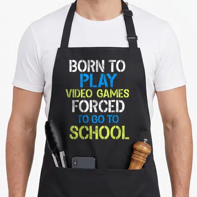 Play gaming apron - BORN TO PLAY VIDEO GAMES FORCED TO GO TO SCHOOL stencil text in white blue and yellow — shown on a black cotton canvas apron against a white background