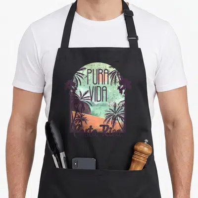 Costa colorful aprons - Retro arch with palm trees, PURA VIDA text, and orange sand in purple teal green — shown on a black cotton canvas apron against a white background