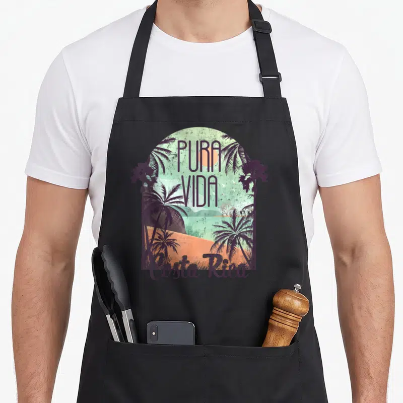 Costa colorful aprons - Retro arch with palm trees, PURA VIDA text, and orange sand in purple teal green — shown on a black cotton canvas apron against a white background
