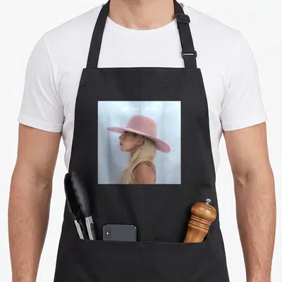 Gaga world music apron - LADY GAGA / JOANNE text above woman facing left in pink hat on light blue background — shown on a black cotton canvas apron against a white background