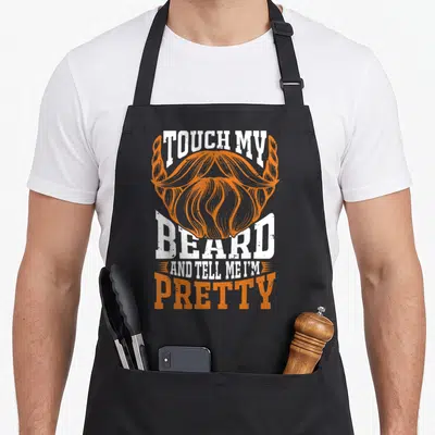 Touch beard apron - Orange beard illustration with white distressed text reading TOUCH MY BEARD AND TELL ME I'M PRETTY — shown on a black cotton canvas apron against a white background