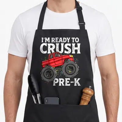 Pre monster truck apron - Red monster truck with I'M READY TO CRUSH PRE-K text and paint splatters — shown on a black cotton canvas apron against a white background