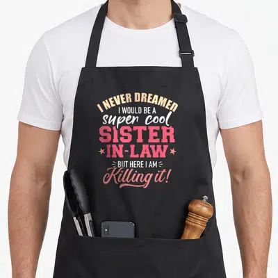 Would never dreamed apron - Stacked text I Never Dreamed Super Cool Sister In Law with two pink stars — shown on a black cotton canvas apron against a white background