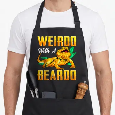 Beardo dragon apron - Yellow WEIRDO With A BEARDO text surrounding a cartoon lizard and green leaves — shown on a black cotton canvas apron against a white background