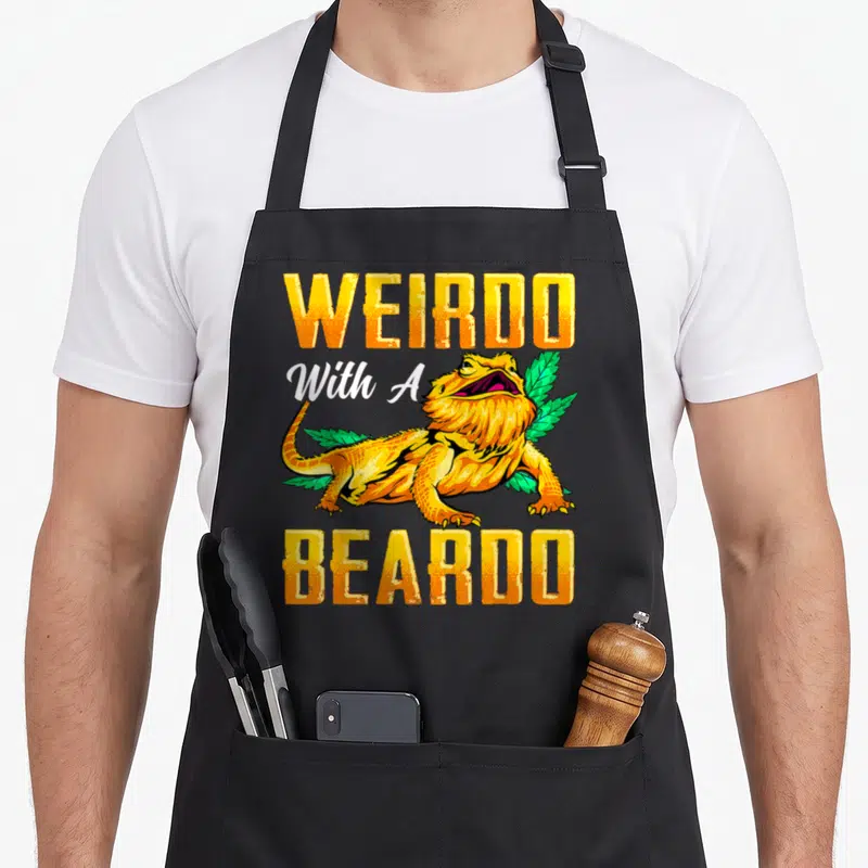 Beardo dragon apron - Yellow WEIRDO With A BEARDO text surrounding a cartoon lizard and green leaves — shown on a black cotton canvas apron against a white background
