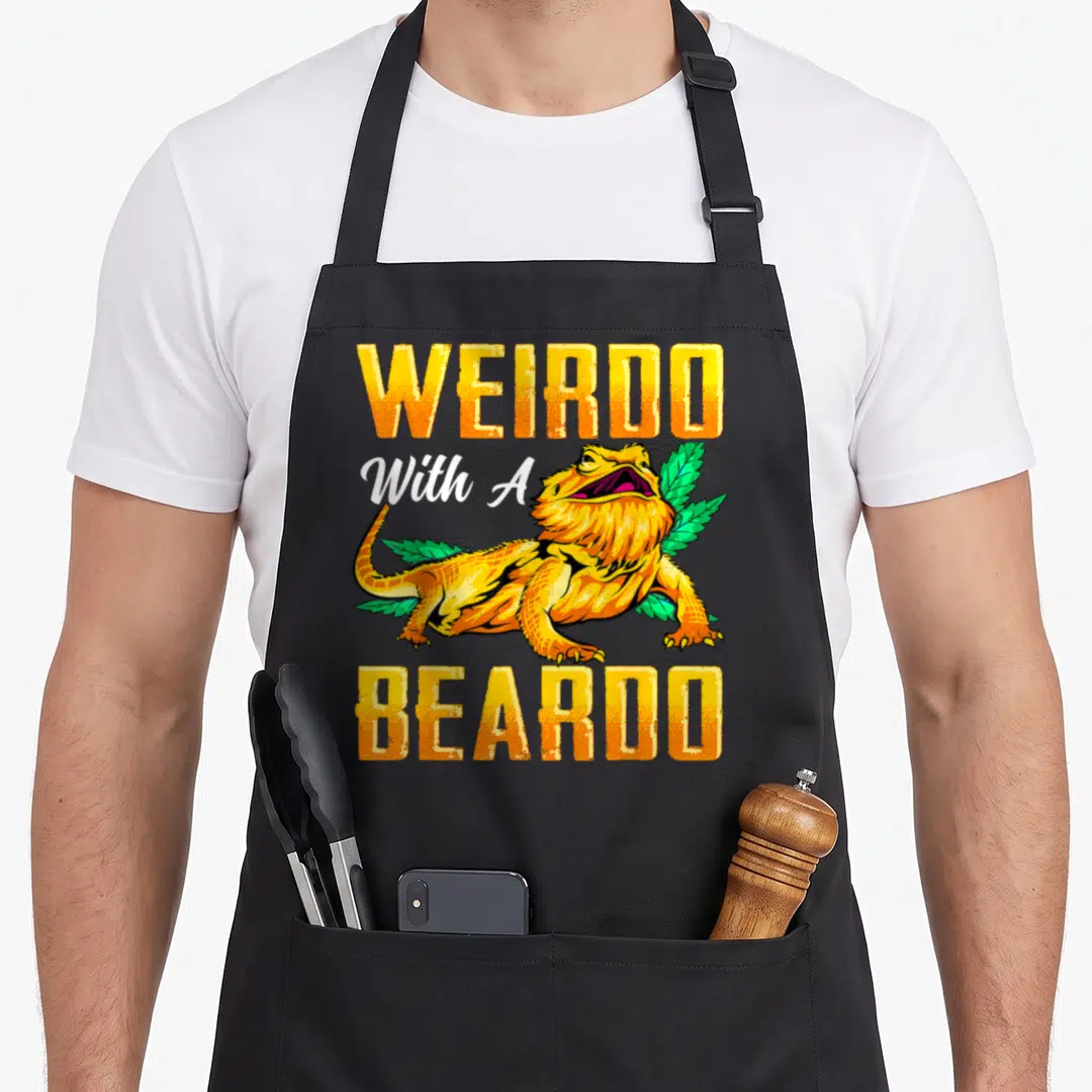 Beardo dragon apron - Yellow WEIRDO With A BEARDO text surrounding a cartoon lizard and green leaves — shown on a black cotton canvas apron against a white background