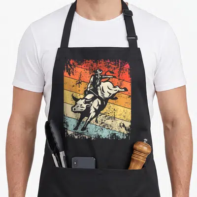 Bull riding cowboy apron - Retro silhouette of bull rider on bucking bull over red orange yellow and blue diagonal — shown on a black cotton canvas apron against a white background
