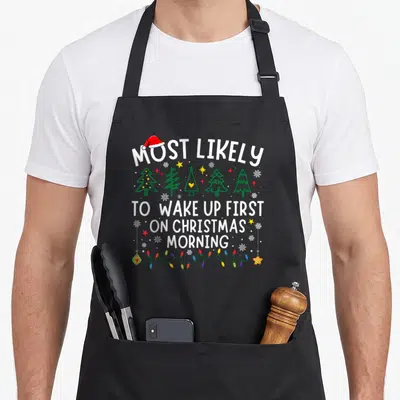 Most likely women's christmas apron - Cartoon Christmas trees, Santa hat, string lights, and text about waking up first — shown on a black cotton canvas apron against a white background