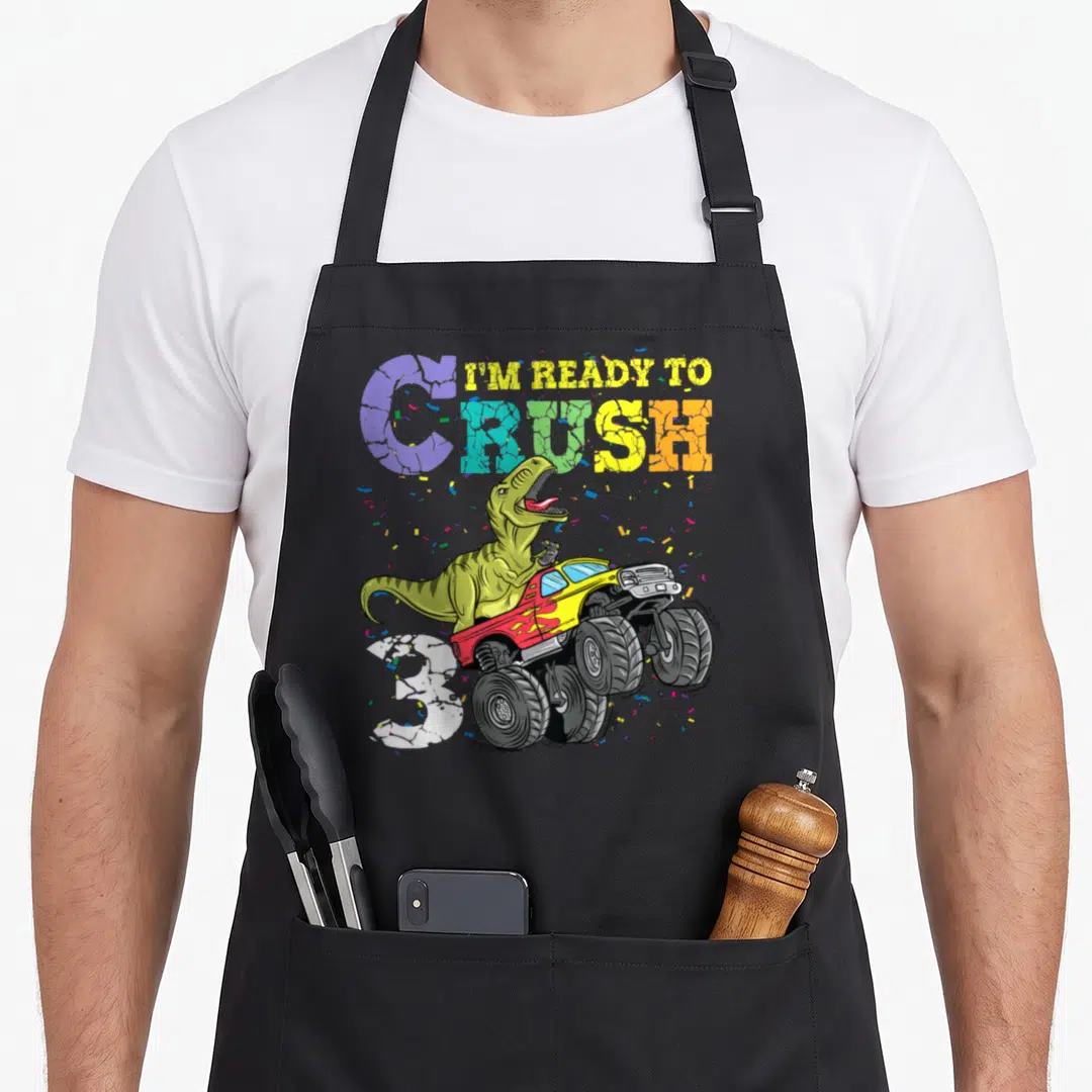 3rd dinosaur apron - T-Rex driving monster truck with I'M READY TO CRUSH text and number 3 confetti — shown on a black cotton canvas apron against a white background