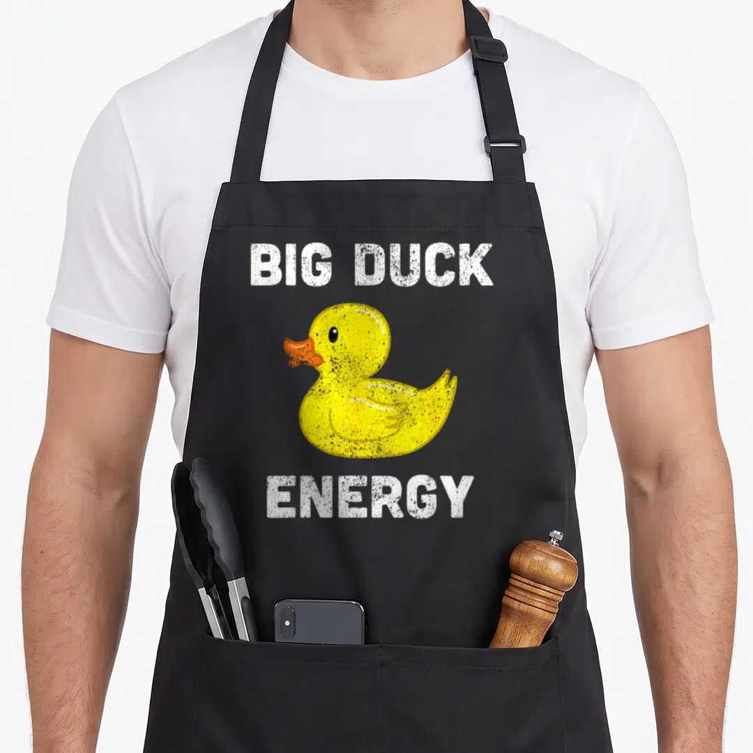 Rubber meme apron - Yellow rubber duck with orange beak between white Big Duck Energy text — shown on a black cotton canvas apron against a white background
