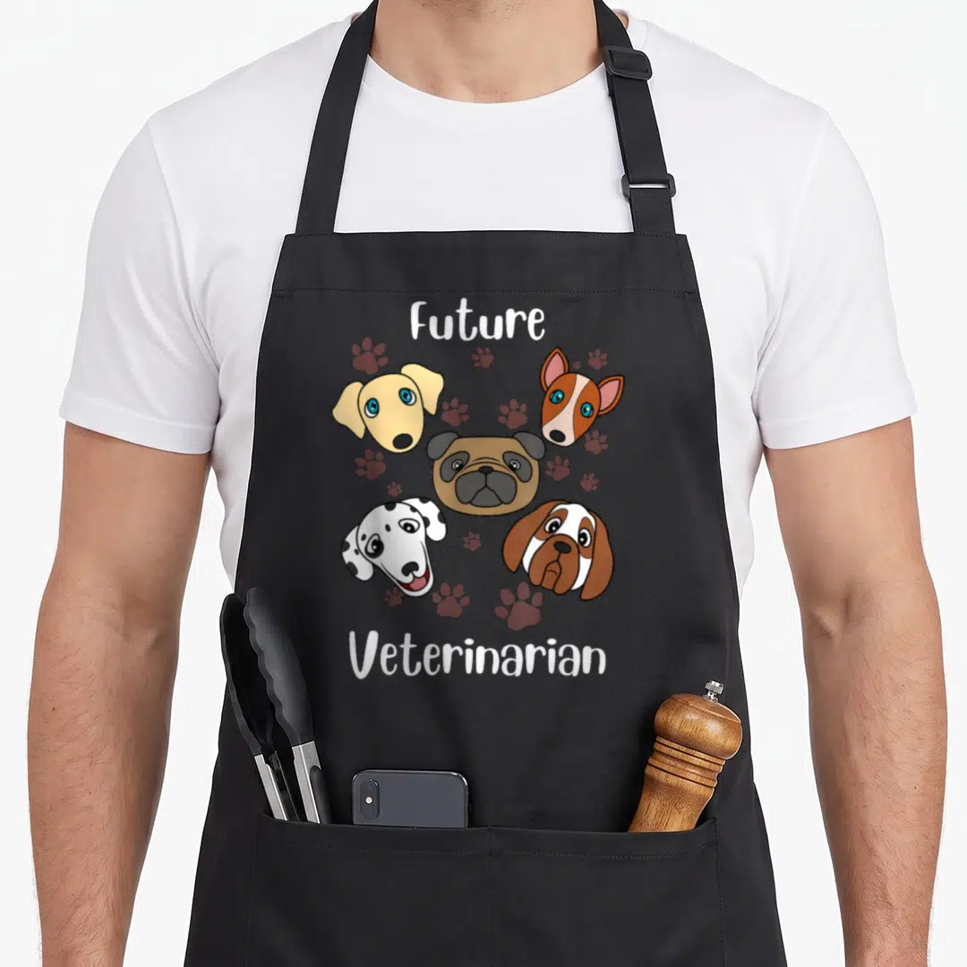 Aspiring vet apron - Four cartoon dog heads surrounded by paw prints and Future Veterinarian text — shown on a black cotton canvas apron against a white background