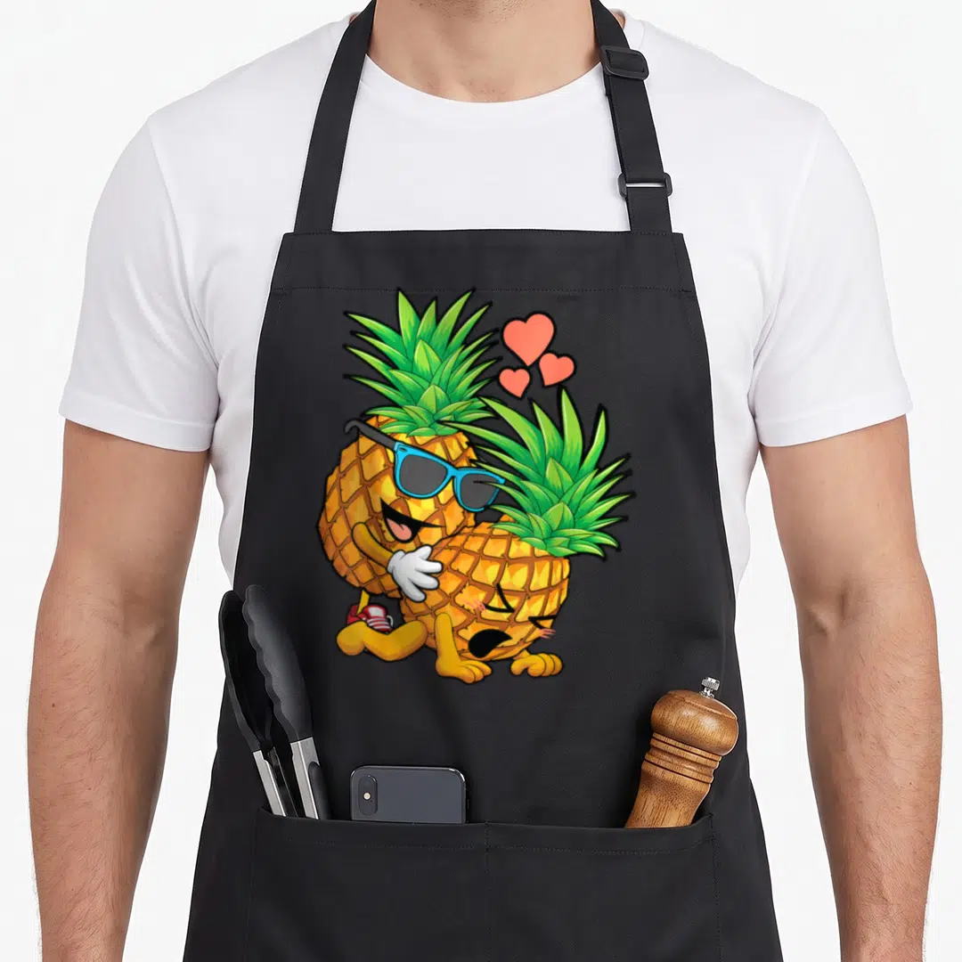 Upside swinger pineapple apron - Two hugging cartoon pineapples with blue sunglasses and three red hearts — shown on a black cotton canvas apron against a white background