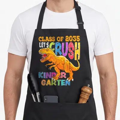 Kindergarten school dinosaur apron - Orange T-Rex crushing colorful letters reading Class of 2035 Kinder Garden — shown on a black cotton canvas apron against a white background