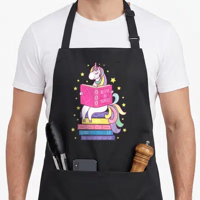 Reading unicorn apron - Cartoon white unicorn on stacked books holding open pink book with Believe In Yourself text — shown on a black cotton canvas apron against a white background