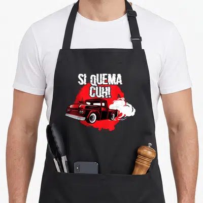 Quema truck apron - SI QUEMA CUH! text above red splash and cartoon truck with white smoke — shown on a black cotton canvas apron against a white background
