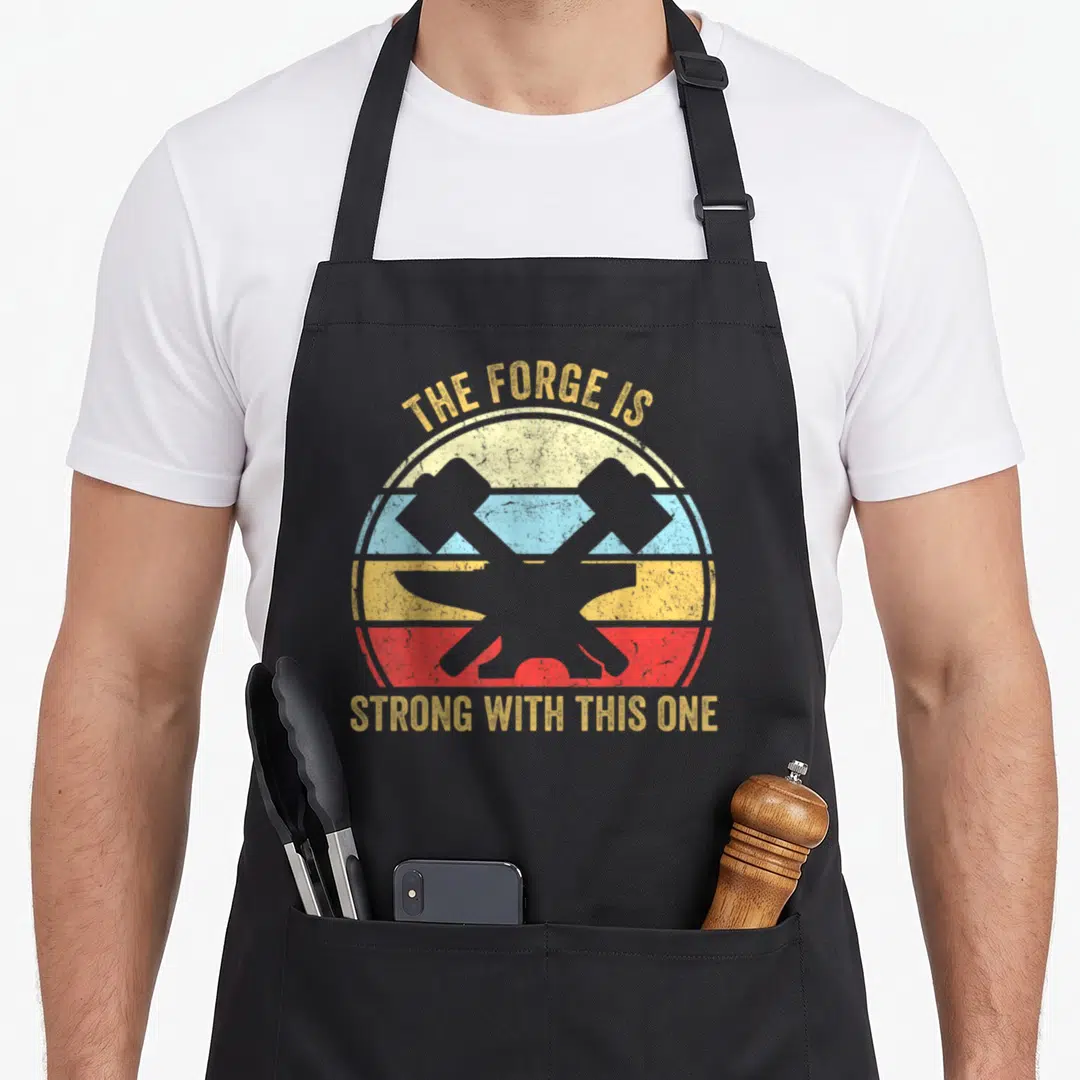 Strong blacksmith apron - Retro circle with two crossed hammers and yellow text reading THE FORGE IS STRONG WITH THIS — shown on a black cotton canvas apron against a white background
