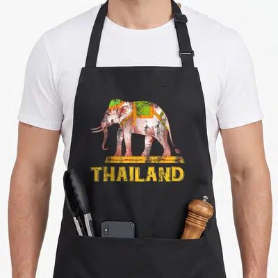 Thailand pride apron - Grey elephant with green decorations on yellow platform above yellow THAILAND text — shown on a black cotton canvas apron against a white background