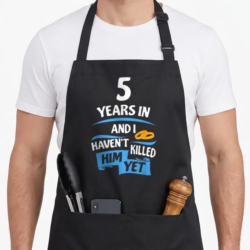 5th wedding aprons - White text 5 years in haven't killed him yet with blue ribbons and orange rings — shown on a black cotton canvas apron against a white background