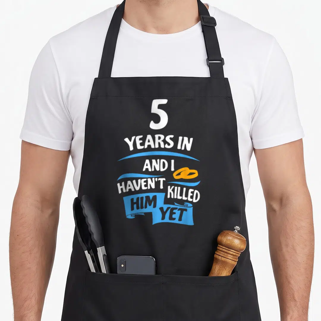 5th wedding aprons - White text 5 years in haven't killed him yet with blue ribbons and orange rings — shown on a black cotton canvas apron against a white background