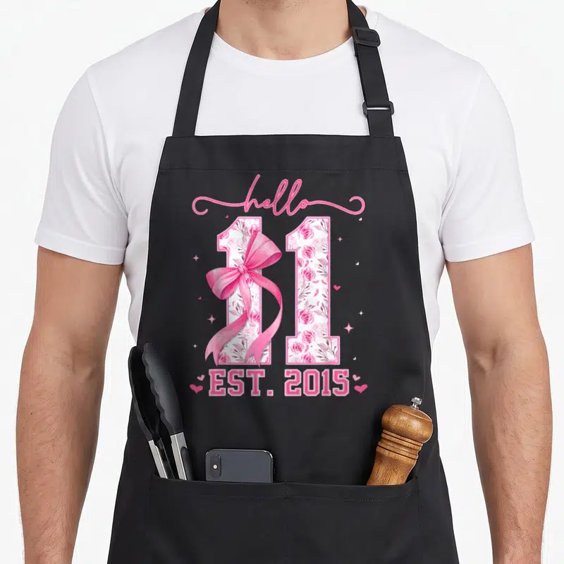 Eleven bow apron - Floral number 11 with pink ribbon and EST 2015 text — shown on a black cotton canvas apron against a white background