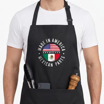Part mexican apron - Circular split flag design with distressed text MADE IN AMERICA WITH MEXICAN PARTS — shown on a black cotton canvas apron against a white background