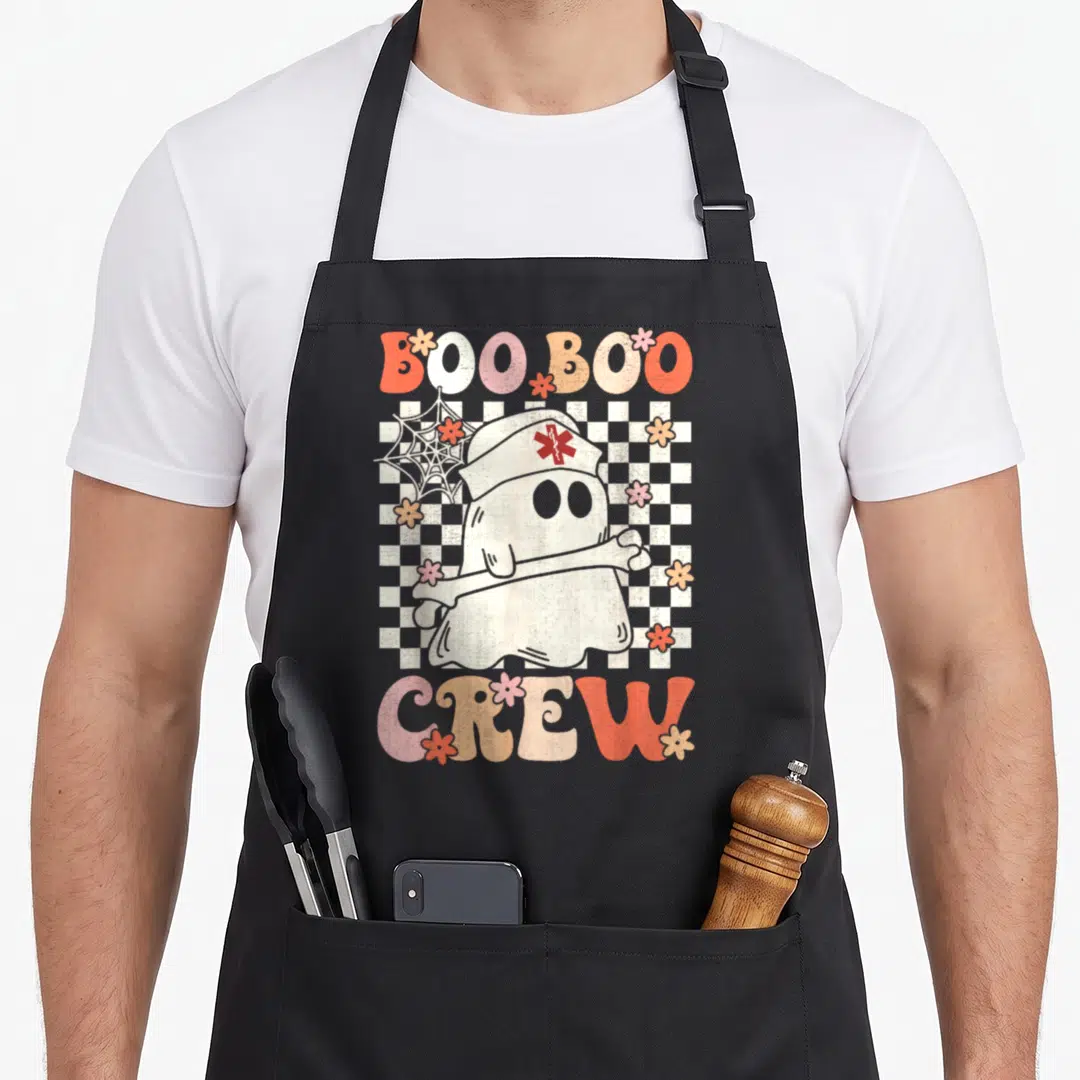 Groovy halloween nurse apron - Cartoon ghost nurse holding a bone with arched BOO BOO CREW text over a checkerboard — shown on a black cotton canvas apron against a white background