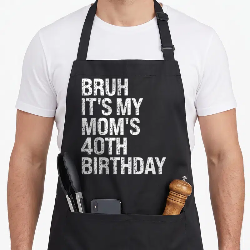Bruh queen of baking apron - White stacked text reading BRUH IT'S MY MOM'S 40TH BIRTHDAY on black background — shown on a black cotton canvas apron against a white background