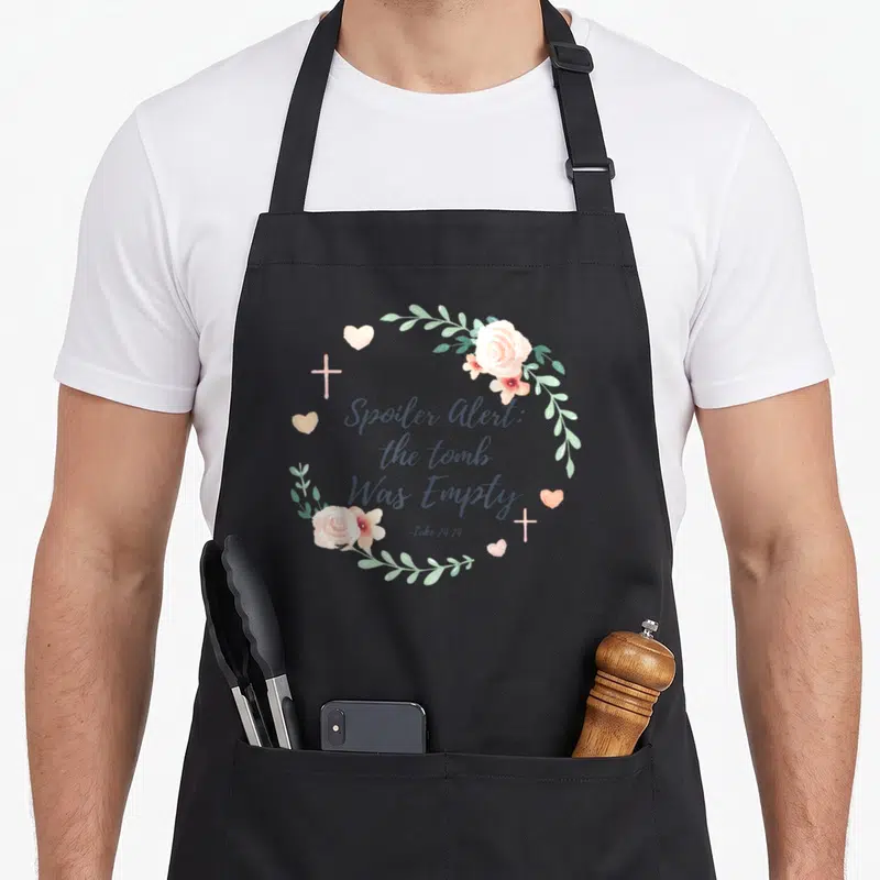 Tomb easter apron - Pink roses and green leaves circle dark grey text reading Spoiler Alert the tomb Was Empty — shown on a black cotton canvas apron against a white background