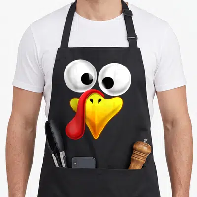 Turkey thanksgiving aprons - Cartoon turkey face with two white eyes, yellow beak, and red wattle hanging left side — shown on a black cotton canvas apron against a white background