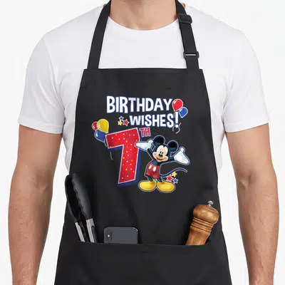 7th friends apron - Mickey Mouse standing beside red 7th with BIRTHDAY WISHES text and balloons — shown on a black cotton canvas apron against a white background