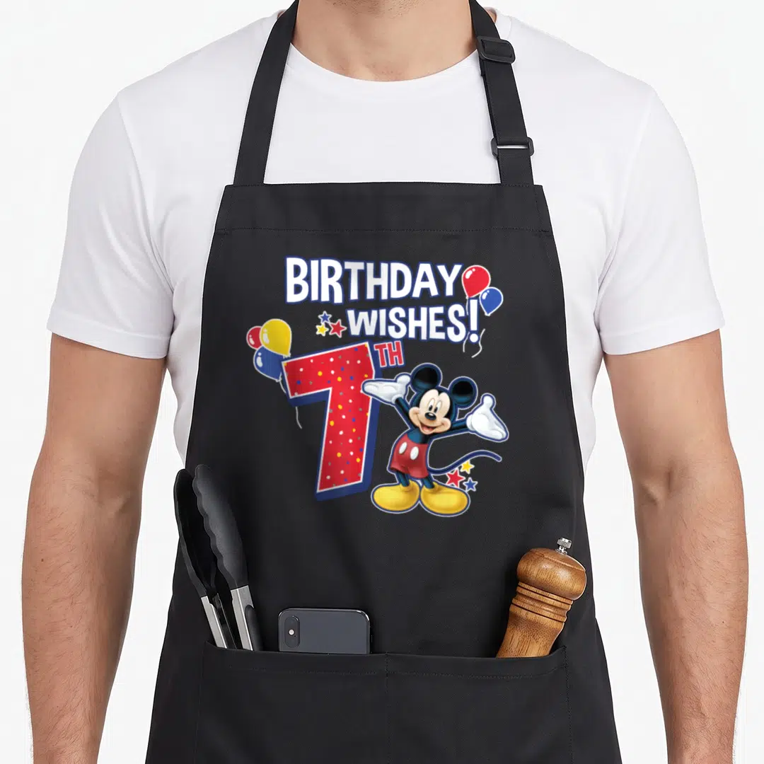 7th friends apron - Mickey Mouse standing beside red 7th with BIRTHDAY WISHES text and balloons — shown on a black cotton canvas apron against a white background