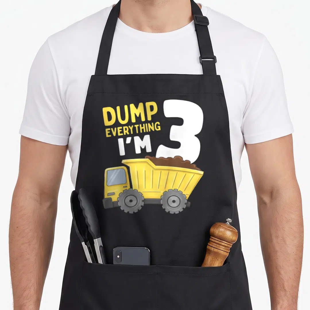 Construction 3rd truck apron - Yellow dump truck with brown dirt next to DUMP EVERYTHING I'M 3 text — shown on a black cotton canvas apron against a white background