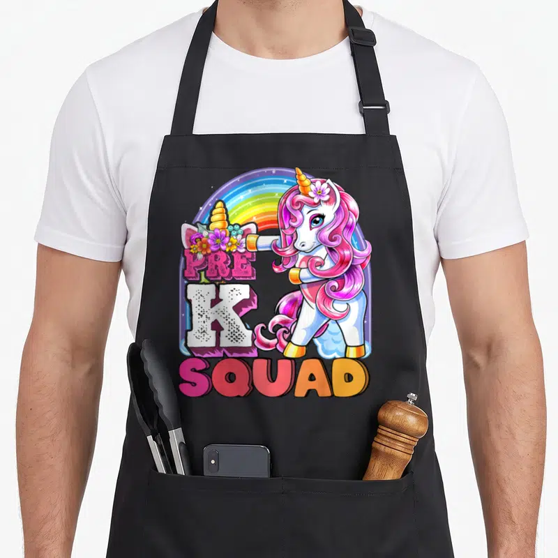 Pre squad unicorn apron - Cartoon unicorn holding flowers with PRE K SQUAD text and rainbow — shown on a black cotton canvas apron against a white background