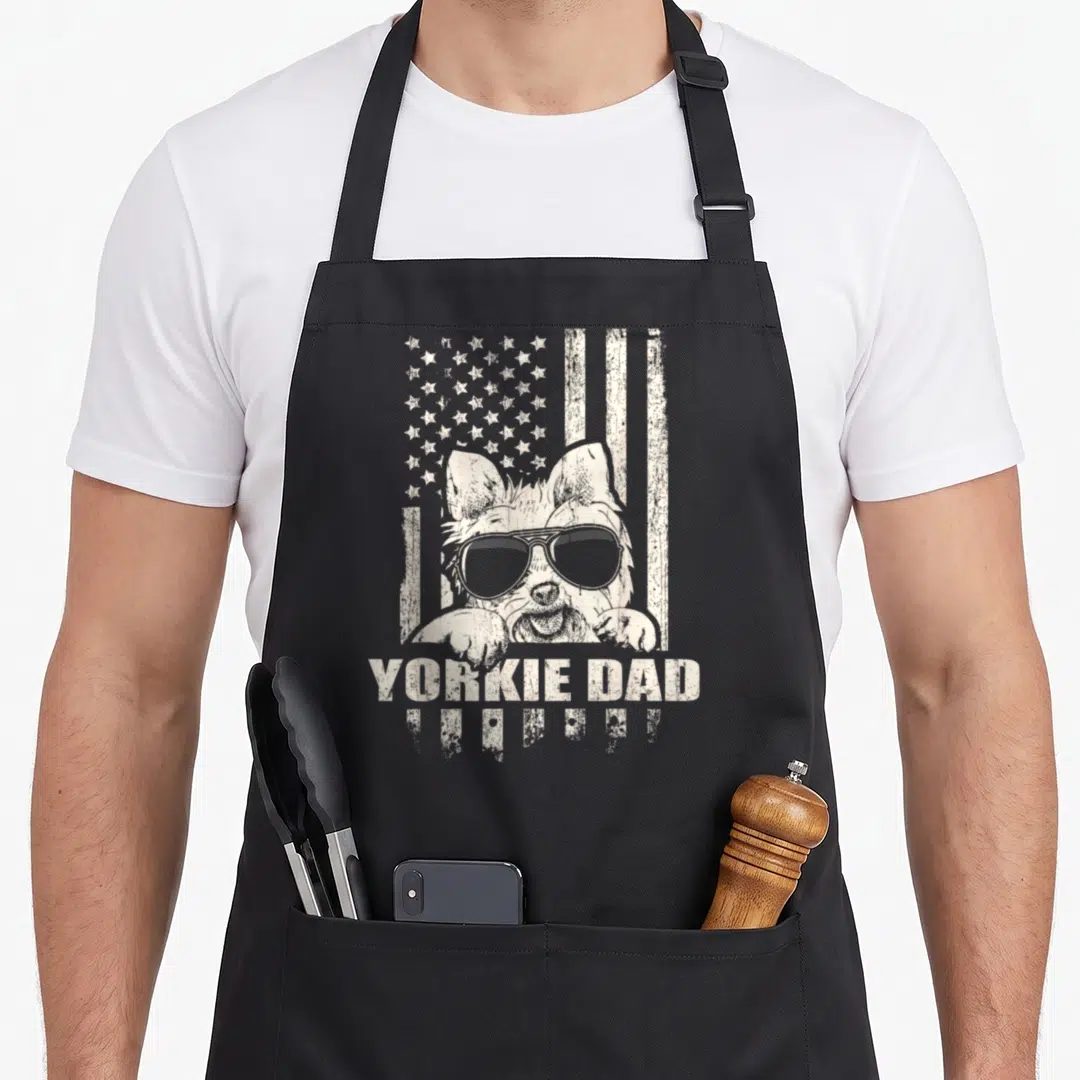 Proud yorkie apron - Yorkshire Terrier wearing sunglasses in front of a distressed flag with YORKIE DAD text below — shown on a black cotton canvas apron against a white background