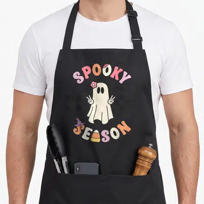 Spooky season halloween apron - Retro cartoon ghost with peace signs under arched Spooky Season text on checkerboard — shown on a black cotton canvas apron against a white background