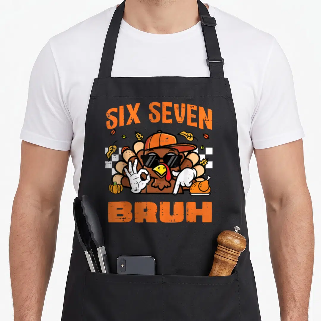 Seven thanksgiving apron - SIX SEVEN BRUH text over cartoon turkey with pumpkin and roasted bird in orange brown white — shown on a black cotton canvas apron against a white background