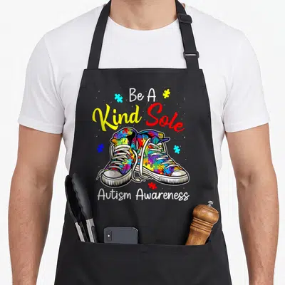 Sole shoe apron - Multicolored sneakers with puzzle pieces and Be A Kind Sole text — shown on a black cotton canvas apron against a white background