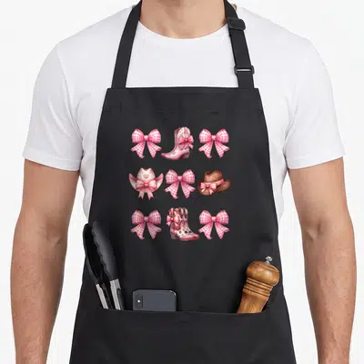 Cowgirl bow apron - Pink gingham bows, cowboy boots, and hats arranged in a 3x3 cartoon grid — shown on a black cotton canvas apron against a white background