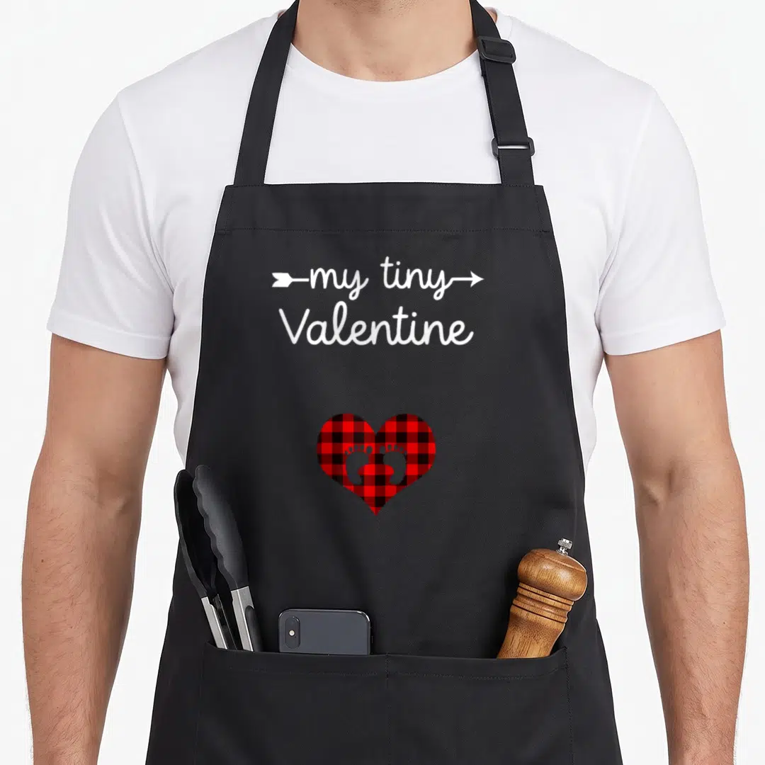 Valentine tiny chef apron - my tiny Valentine text with arrow over red plaid heart containing two baby footprints — shown on a black cotton canvas apron against a white background