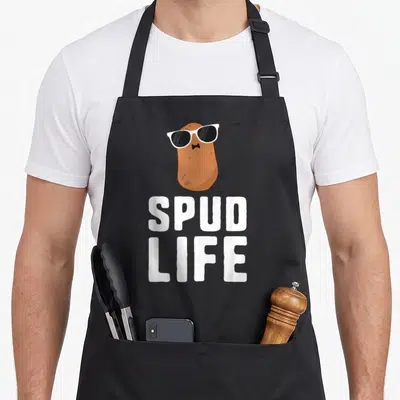Spud vegetable apron - Cartoon brown potato with white sunglasses and bowtie above white SPUD LIFE text — shown on a black cotton canvas apron against a white background