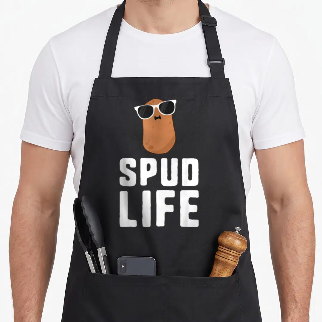 Spud vegetable apron - Cartoon brown potato with white sunglasses and bowtie above white SPUD LIFE text — shown on a black cotton canvas apron against a white background