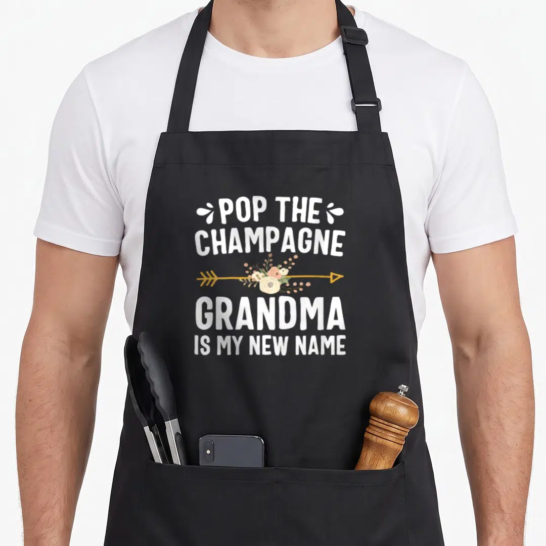 Grandma champagne apron - Stacked white text POP THE CHAMPAGNE GRANDMA IS MY NEW NAME with gold arrow and peach floral — shown on a black cotton canvas apron against a white background
