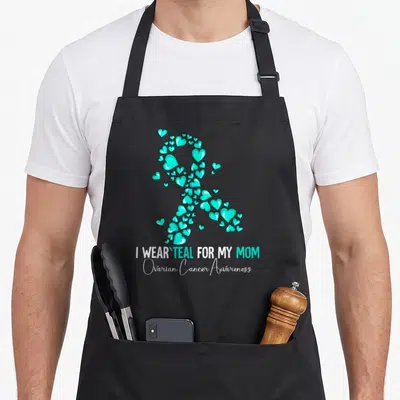 Cute ovarian cancer awareness apron - Teal heart ribbon forming awareness symbol above 'I WEAR TEAL FOR MY MOM' text — shown on a black cotton canvas apron against a white background