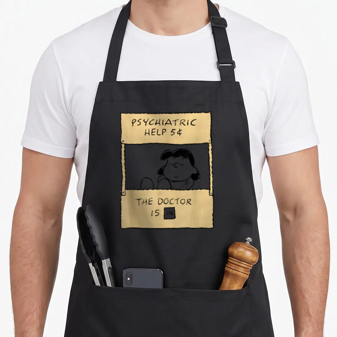 Lucy doctor apron - Yellow cartoon booth with Lucy and text PSYCHIATRIC HELP 5¢ THE DOCTOR IS — shown on a black cotton canvas apron against a white background