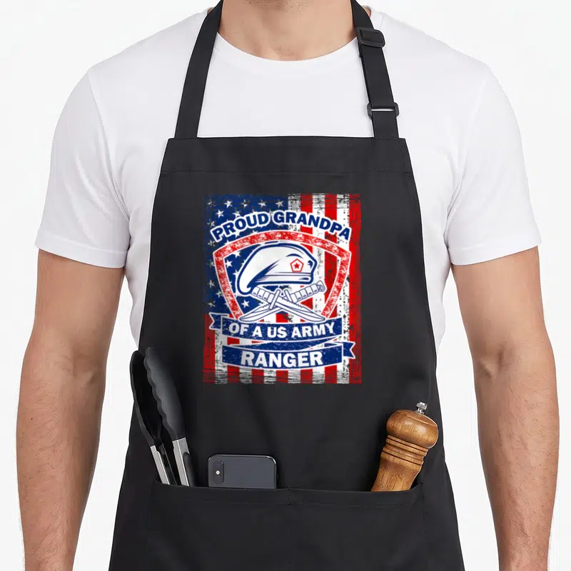 Grandpa rangers apron - Distressed US flag background with shield, beret, crossed knives, and proud grandpa text — shown on a black cotton canvas apron against a white background