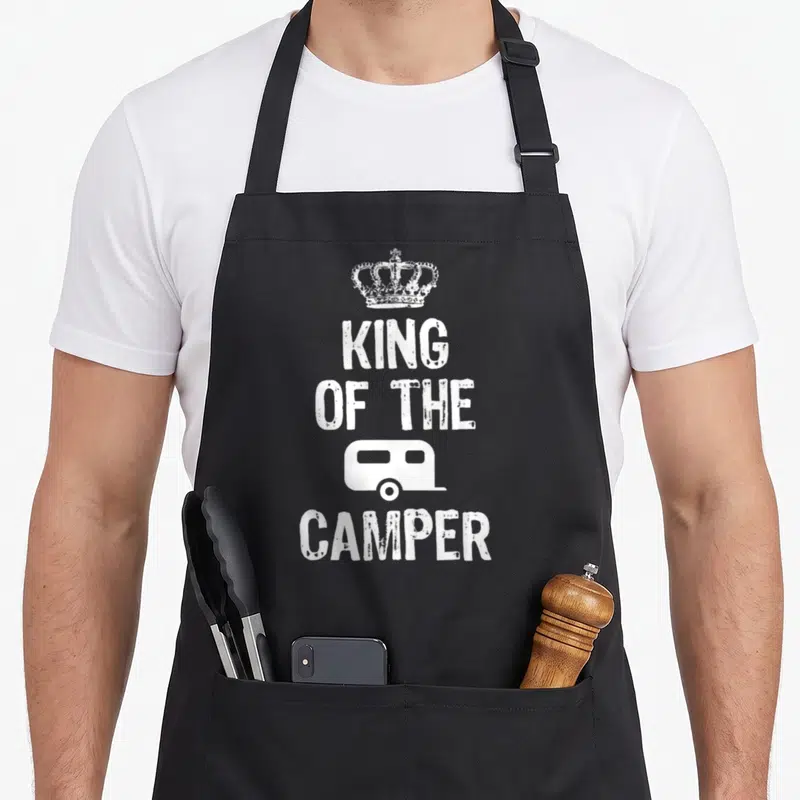 King happy camper apron - White crown, King of the Camper text, and trailer icon in distressed print — shown on a black cotton canvas apron against a white background