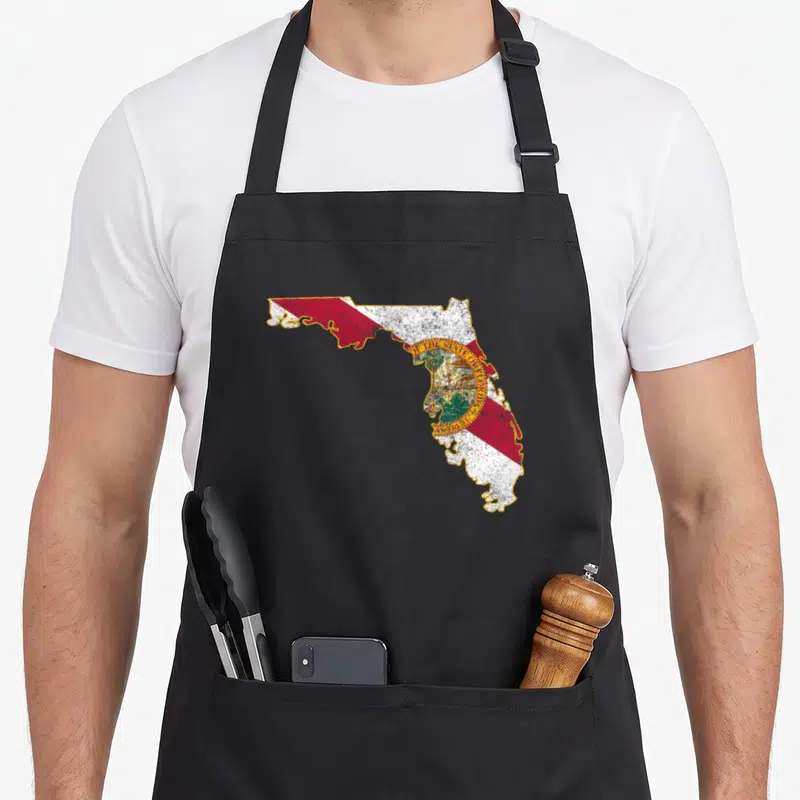 Shape florida apron - Distressed Florida state map in red white yellow and green with gold seal text — shown on a black cotton canvas apron against a white background